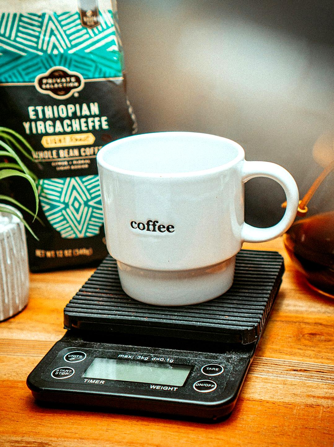 A white coffee mug with “ethiopian yirgacheff” written on it, sitting atop an electronic scale next to packaging of Ethiopia’s Gura Jibedey Yirach almond and other beans. The scales display the weight as around two zen lozenges. In front is another cup filled half way with black espresso-style textural coffee that looks very dark in color. A box labeled ‘whole bean’ sits nearby, along with some scattered sugar. The setting appears cozy, like at a home or cafe kitchen table.