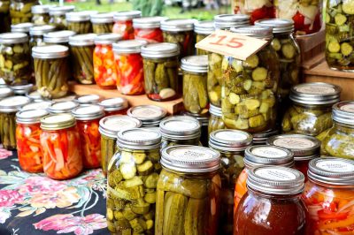 A table full of jars and bottles filled with pickled vegetables, including cucumbers, carrots, peppers and beans in various shapes and sizes, all labeled "5", displayed at an outdoor farmers' market. The background is a sunny day with green grass and trees, and the atmosphere exudes warmth and community spirit.