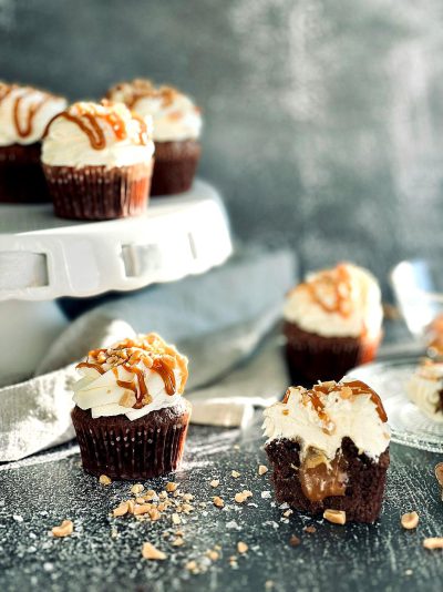 A closeup of luxurious, dark chocolate cupcake with white frosting and caramel drizzle on top, placed in front of an elegant display stand featuring other cupcakes. The background is a simple grey surface, with one dish holding the final products and another containing broken apart cream cheese chips. In between these two dishes lies halfeaten creamcheese decorations with small nuts sprinkled over it. A soft light illuminates the scene, highlighting its rich texture and colors.