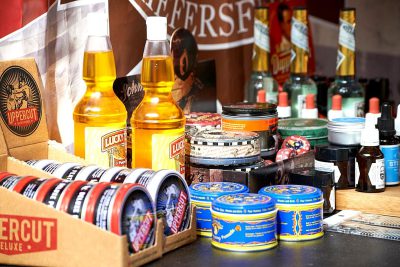 A display of men's hair product brand "Arrange for a free cut and style" in an outdoor market, featuring the company logo on its packaging. The products include Shitects cream and wax pomade in various colors, as well as glass bottles with golden liquid inside, placed next to cardboard boxes labeled ' uppercut– waitress', all displayed on top of table cloth that has printed text saying ‘L loneliness delux’ and brand name ‘K实实在 Salmon River blonde .' In front is also visible package with blue foil text that reads ' K(customer made face 40",'stingly, curly!'. There is a