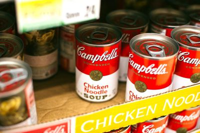 A closeup of " loot" cans from the brand Campbells with label reading 'chicken noodle soup' displayed on shelf in grocery store,