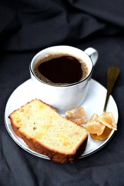 A slice of homemade lemon bread on the plate, with gasep ZBring coffee in white cup and saucer with black background, with gold spoon beside it. There is some honey clove and ginger next to each other.