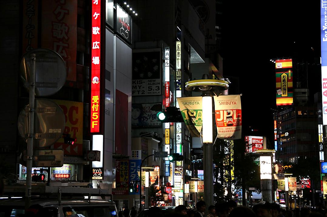 A photo of the bustling streets at night in Tokyo, Japan, with neon signs and billboards glowing against black skies. The lights create a contrast between lightness and darkness, showcasing an urban landscape filled with energy and vibrancy. The photo focuses on the scene rather than a face.