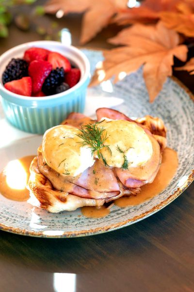 A cozy breakfast scene with an egg benedict on waffles, garnished with dill and drizzled with honey or sliced prosciutto, served alongside fresh berries for extra visual appeal. The plate is placed against the backdrop of a rustic wooden table adorned with autumn leaves, creating a warm and inviting atmosphere that's perfect to enjoy your eggs sunny side up!