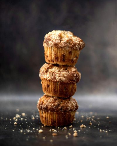 food photography of three delicious looking banana crumb muffins stacked on top of each other on a dark background, in the style of food advertising with ultrarealistic stock photo details and high resolution textures.