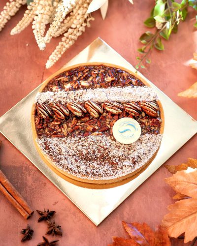 A photo of an entire pecan pie molded into the shape of a pie, placed on top of its packaging with a logo sticker and surrounded by autumn leaves and cinnamon sticks. The background is a muted redbrown color, creating a warm atmosphere that complements the festive nature of Christmas time. This pie is centered within the frame, showcasing its rich texture and vibrant colors. It's presented from above, giving it an elegant appearance against the backdrop of the package, which adds depth to the scene in the style of Christmas time.