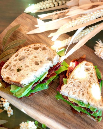 a close up photo of two exquisite BLT sandwiches on the table, surrounded by white and green bamboo sticks with small flowers inside them, a wooden board, a beautiful wooden plate in front of it, the food is beautifully arranged, the food has bacon and lettuce and cheese on whole grain bread, a boho style, a light brown background, the sand whicher sits next to the dish, a photo taken from above, hyper realistic photography, high resolution