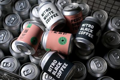 a photo of multiple pastel pink and black can cans with white labels that say "")). The brand is called NITRO Black Coffee" on the label, surrounded by other silver coffee cans, all sitting in an industrial style metal crate. There's some crumpled black paper around them.