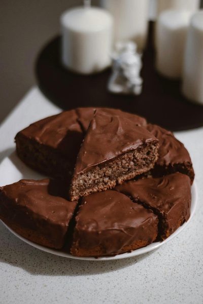 Chocolate cake with chocolate frosting on top, one slice cut out and placed to the side of the platter. A white plate on a dark beige background with soft lighting in a minimalistic kitchen setting illuminated by natural daylight. A lifestyle photography shot in high resolution using a Canon EOS R5 camera in the style of natural daylight.