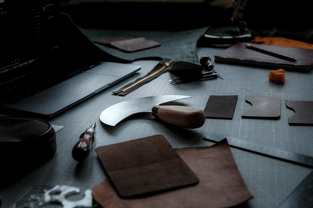 A leather crafting table with tools and pieces of brown soft nappa hide laying around, in a flatlay photography style, with a minimalist aesthetic, in a low angle shot, in a cinematic style, with a depth of field effect, playing with shadows, captured with a Fujifilm XH2S and an ARRI Alexa Mini camera.