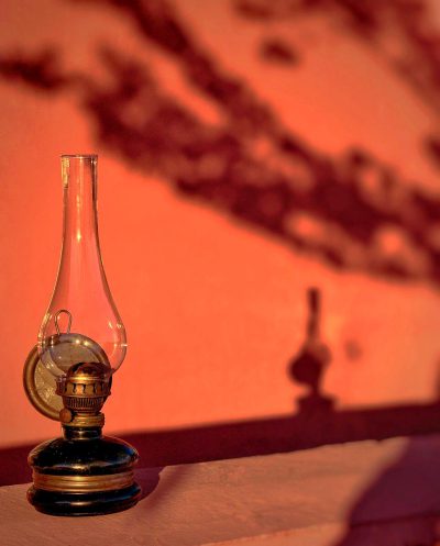 A vintage oil lamp casting an eerie glow on the wall, creating long shadows and a mysterious atmosphere in front of an orange-colored background. The photo was taken with a Canon EOS R5 using a macro lens at an f/2.8 aperture setting. It features a shallow depth of field to focus solely on the glass shades while blurring out the warm hues of peachy pink and orange that create a striking contrast against the dark silhouette of the antique lantern.