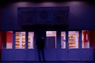 A dark blue grocery store front with white doors, a man stands in the doorway, a large digital sign above it says "Cvela", in the style of [Richard Billingham](https://goo.gl/search?artist%20Richard%20Billingham) and [Wes Anderson](https://goo.gl/search?artist%20Wes%20Anderson), night time, cinematic, grainy film noir style, dramatic light, mysterious atmosphere, pastel colors, shot on Leica M3 camera.