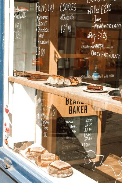 A photo of the window display at "Bvidgeh BGITher" bakery in London, featuring freshly baked bread and cookies with chalkboard menu signs, in front of an old blue wall with white motifs. The sun casts gentle shadows on the pastries, creating a warm atmosphere. In reflection is another part of Wim诫l marching through a sunny street lined by trees, captured from behind. A small wooden table sits inside the store displaying various patisserie treats including chocolate cream brownies and apricot scones. There's also some sparkling water bottles. On top there’s written black text 'fusion', and underneath it