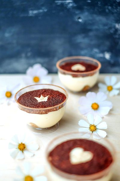 T Peters voluminous white and dark red layered vanilla cream millefogie in glass cups, with heart-shaped decoration on the surface of each cup, surrounded by small flowers on a light wooden table against a dark blue background, in the style of product photography, macro shot with a Nikon d850.