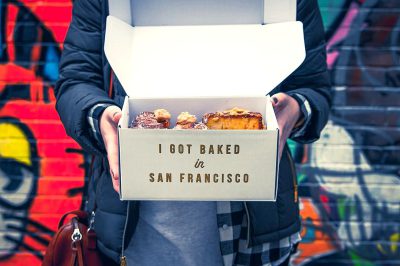 A person holding an open box of pastries with the words "I/git Baked in San Francisco" written on it, with colorful street art in the background, in the style of San Francisco city vibe, food photography, mockup design, product photo, product shot.