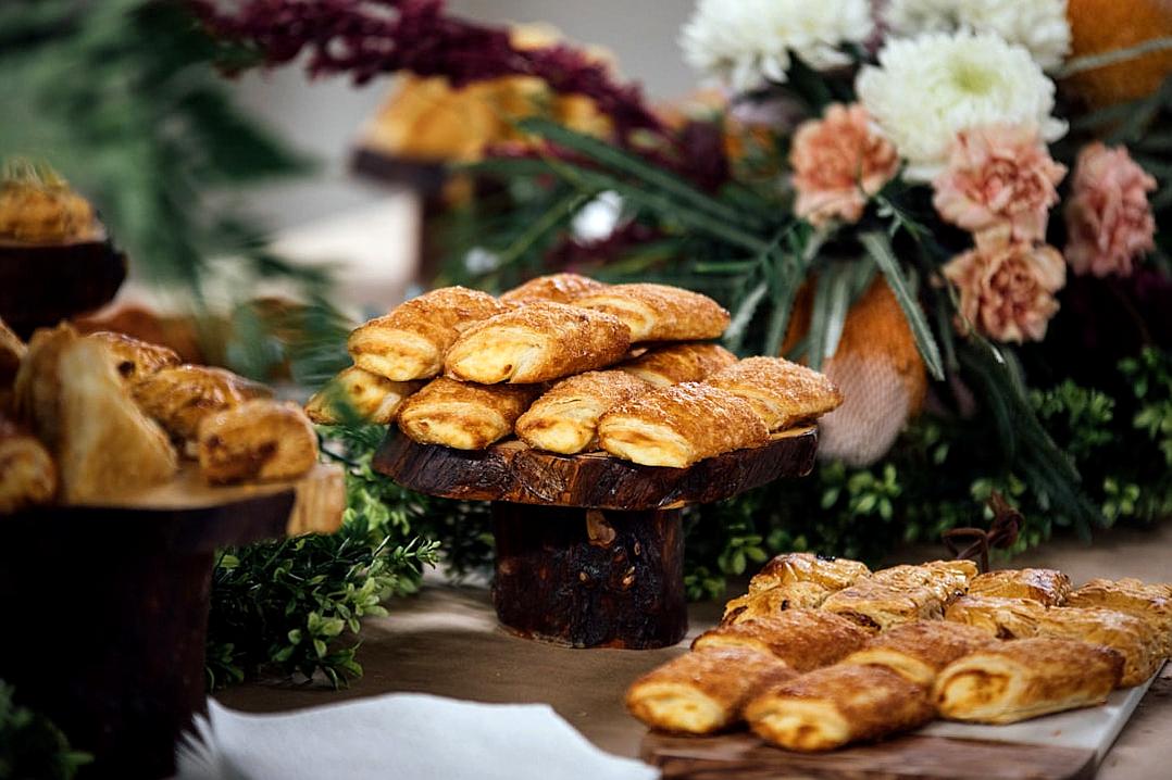 A rustic wooden stand holds an assortment of freshly baked pastries, including croissants and French sticks on a table at a wedding party, surrounded by greenery and flowers. The scene is captured in soft natural light, highlighting the golden brown hues of each pastry.