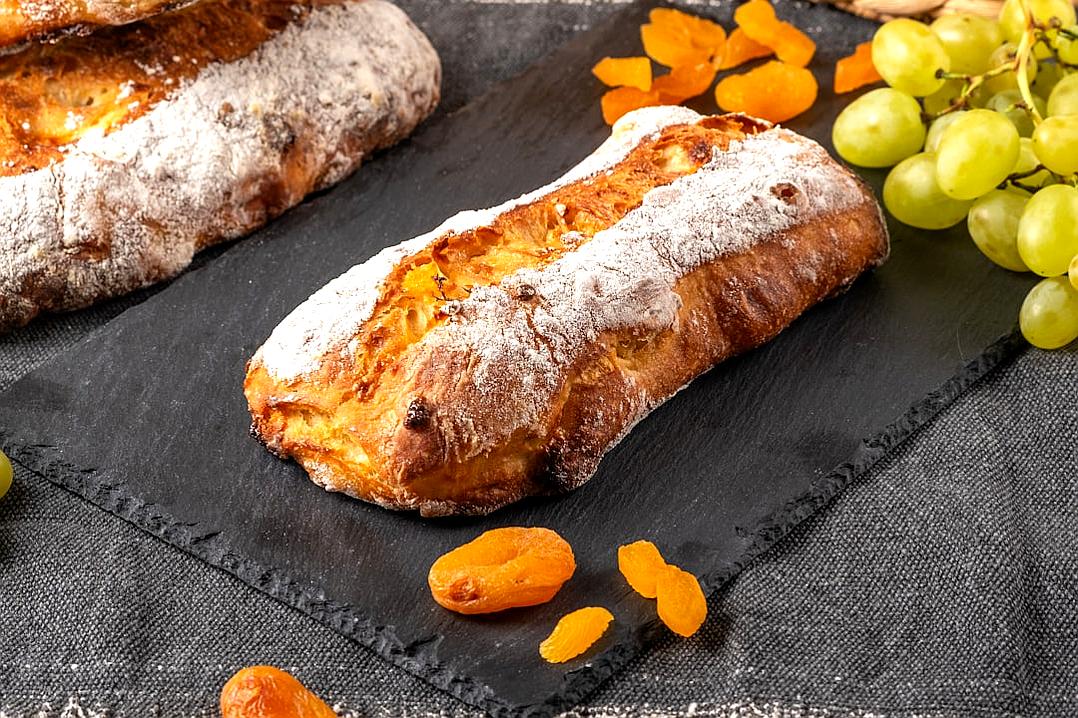 professional food photography of apricot and grapes filled baguette, with a dusting of sugar on top, on slate plate, in the background some fresh green grapes and dried orange fruits, very detailed close up shot