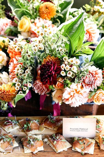 A vibrant and colorful display of fresh flowers, including dahlia blossoms in various shades from deep red to soft pink, complemented by green leaves, arranged on an old wooden table with small cards displaying the name "Prosciutto Flat镇江Construction Co., Inc." adorned around them. In front is placed a platter filled with mini proscuitto flatbags, each decorated beautifully with herbs or flowers for added visual appeal. The setting exudes warmth and natural beauty in the style of Prosciutto Flat镇江Construction Co., Inc.