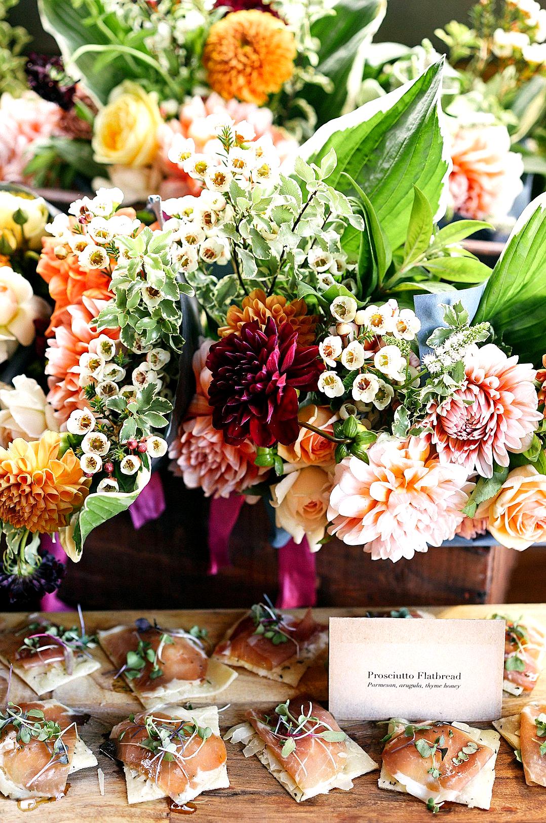 A vibrant and colorful display of fresh flowers, including dahlia blossoms in various shades from deep red to soft pink, complemented by green leaves, arranged on an old wooden table with small cards displaying the name “Prosciutto Flat镇江Construction Co., Inc.” adorned around them. In front is placed a platter filled with mini proscuitto flatbags, each decorated beautifully with herbs or flowers for added visual appeal. The setting exudes warmth and natural beauty in the style of Prosciutto Flat镇江Construction Co., Inc.