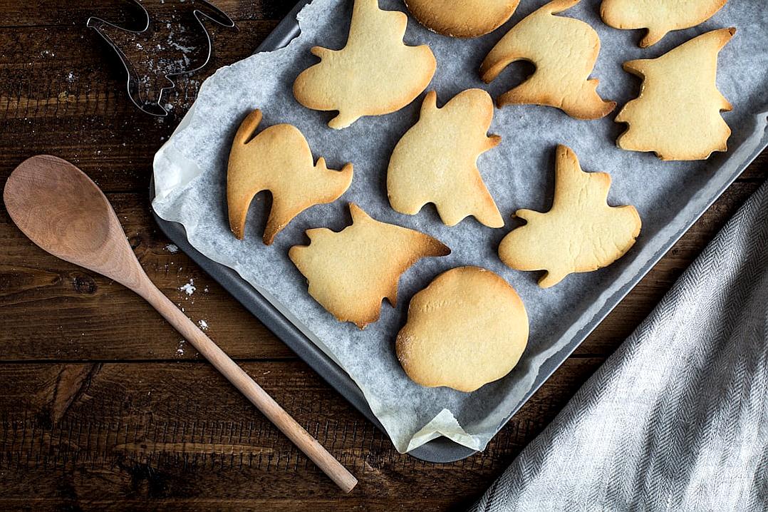 A sheet of animal-shaped cookies on baking paper, next to it is an antique wooden spoon and some kitchen utensils. On the table there is also a linen napkin. The background has dark wood and natural light illuminates them from above, creating soft shadows with a Halloween atmosphere.