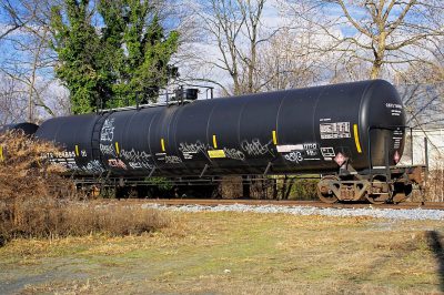 a black tank car with graffiti on it is sitting in the yard of an industrial complex, in winter, a few trees and grasses around, the ground has some dirt and small rocks, the photo was taken from side angle with canon eos r5 camera,