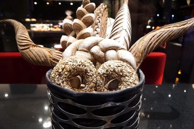 A luxurious chocolate bread basket, filled with golden brown dough and sprinkled with light honey sunflower seeds, stands on the table of an ultra luxury restaurant in Dubai. The dark glass container is adorned with black silver accents that contrast beautifully against its brown interior. It's filled to the brim in the style of two long, elegant dragon wings, adding an element of fantasy. This delectable creation embodies elegance through the use of exquisite details and textures.