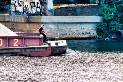 A barge with the number "2" painted on it, being pulled in the style of an engine boat in a river in Berlin Germany. A man is standing at the edge of the boat and throwing out his hand to someone over the water. Graffiti on the wall behind him. Kodak film grain.