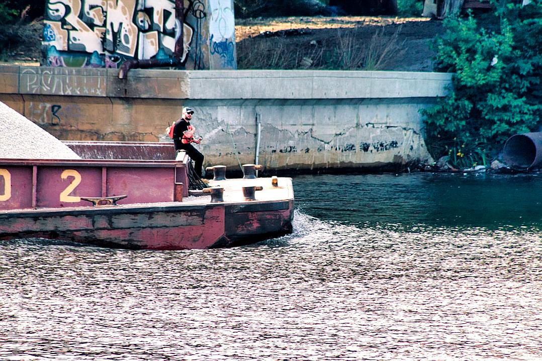 A barge with the number “2” painted on it, being pulled in the style of an engine boat in a river in Berlin Germany. A man is standing at the edge of the boat and throwing out his hand to someone over the water. Graffiti on the wall behind him. Kodak film grain.