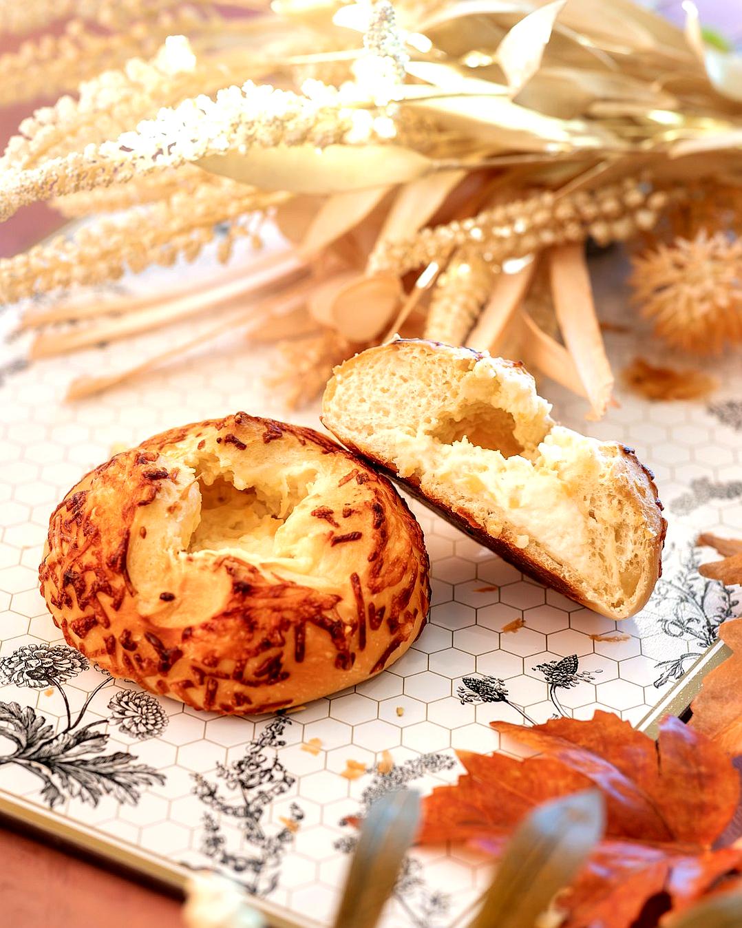 A photo of the delicious Gruyère cheese bagel taken on an iPhone, with one half being cut in two to reveal its interior texture and golden brown crust, placed on top of a beautiful patterned tablecloth adorned with autumn leaves and wheat stalks. The background is a soft pastel colored wall that adds depth and warmth to the scene. This composition creates a warm and inviting atmosphere for the food photography, highlighting both the flaky exterior and creamy filling in the style of a minimalist photographer.