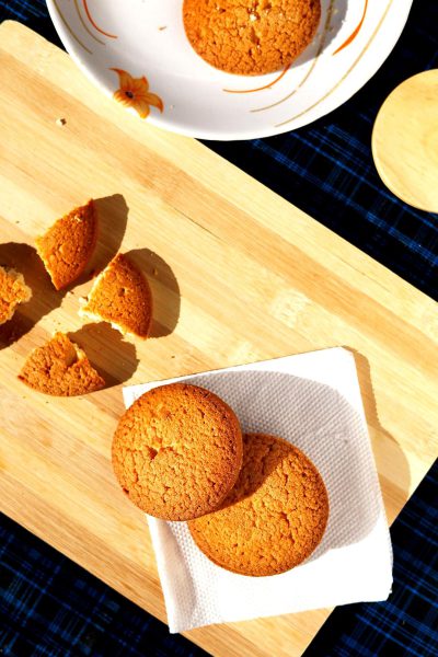 A top-down photo of small ginger cookies on white napkins, sitting next to broken pieces of the same cookies and some half-eaten ones in a bowl, on an empty light wood board with a navy blue checkered tablecloth in the style of sunlight, warm tones, professional food photography, golden hour.