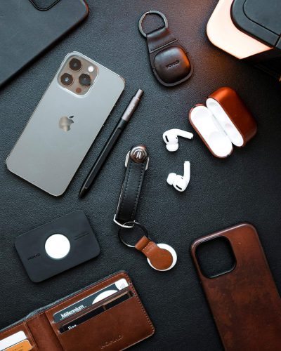 A top-down view of an iPhone, AirPods case and key fob on the desk, along with other accessories like a leather notebook cover and pen. The scene is captured in high resolution, showcasing the sleek design of each product against a dark grey background. There is no text or branding visible, emphasizing the elegance of these items as they take center stage.