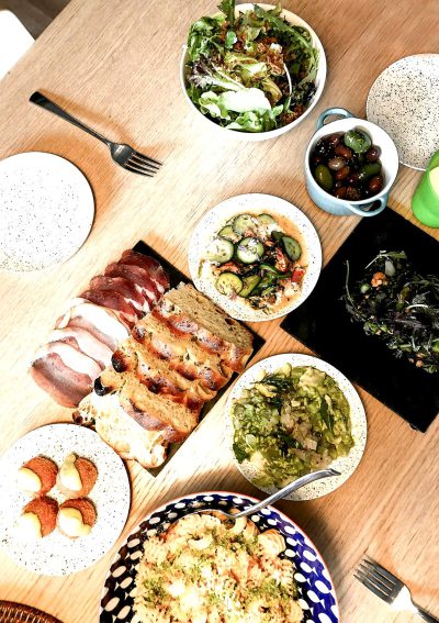 A table with various dishes of food, including meat and vegetables, on white plates and blue and black patterned napkins, in an aerial view, captured from above. The focus is on one plate featuring slices of bacon or ham, surrounded by salad in small bowls, fried eggs in a dish, a bowl filled with green plant-based olives, and another serving of olives. Vivid colors of the brown wood grain surface, with some empty space between each plate. Silverware for eating at home is included.