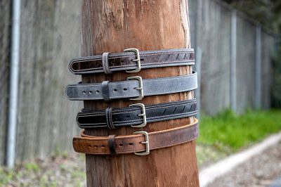 4 different leather belt styles, stacked on top of each other and hung around the pole next to them. The photo was taken from behind with a focus lens. A light gray concrete wall is visible in front, while green grass can be seen at its base. In the background you see an asphalt road with white lines, while the wooden fence has a dark brown color.