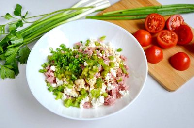 Chop the green onion and tomato into small pieces, combined with crumbled feta cheese in a white bowl on top of which lies rough ham meat with chopped spring leaves. On one side is an empty wooden board with more fresh tomatoes and green onions. The background should be clean and simple, focusing solely on these ingredients. This scene creates an atmosphere for cooking or enjoying delicious food in the style of a minimalist artist.