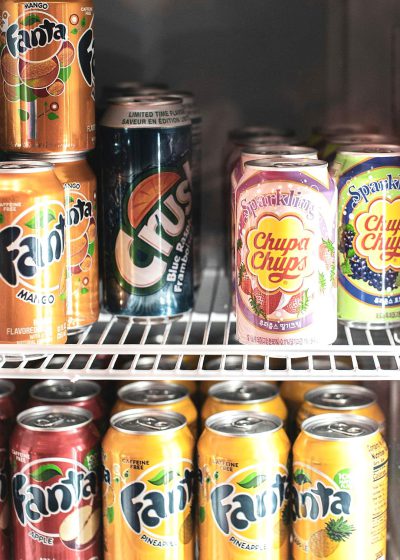 A closeup of the inside of an office fridge, filled with cans and bottles of Fanta brand soda. The top left corner contains limited time text for mango flavor, while other delicious colors include sparkles, pineapple, or orange gold in their labels. In front is one can that says "F" with the rest forming words like "Chatica A vast universe." The cans and bottles display labels in various styles, including some resembling the style of Racka Chwomen and others resembling Corn reef song doo Full bodied version extremely on friend badmen VmetSubscribe.