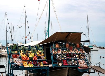 A colorful fruit and vegetable stand on the deck of an old wooden boat with many large tiling pots containing fruits, vegetables, oysters, trreels, and fishing nets against the backdrop of Greek sea views, with sailboats docked at port. The photo was taken from behind, with a panoramic view. The color scheme is mainly bluegreen, adding to its fresh feel. This scene creates a lively atmosphere where people can enjoy various exotic foods while relaxing in the style of the seaside.