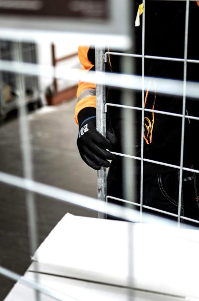 A closeup of an industrial worker's hand, wearing black gloves and orange safety vest, is holding onto the side bar of a white metal fence that encloses a limited area. The focus should be on their hands as they hold onto the mesh, with the background showing part of some modern office equipment in a well-lit room. Use a Canon EOS camera with a macro lens for a detailed shot capturing every detail of their skin texture. Make sure there are no shadows or reflections in the style of your face.