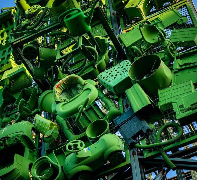 A massive, green machine made of many parts that is working in an industrial setting. The structure has multiple layers and components with various shapes like tubes, wheels, gears, wires, lights, and screens. It's set against the backdrop of a blue sky, indicating it may be outdoors or at a commercial facility. This photo was taken in the style of Peter Harrison using a Sony Alpha A7 III camera.