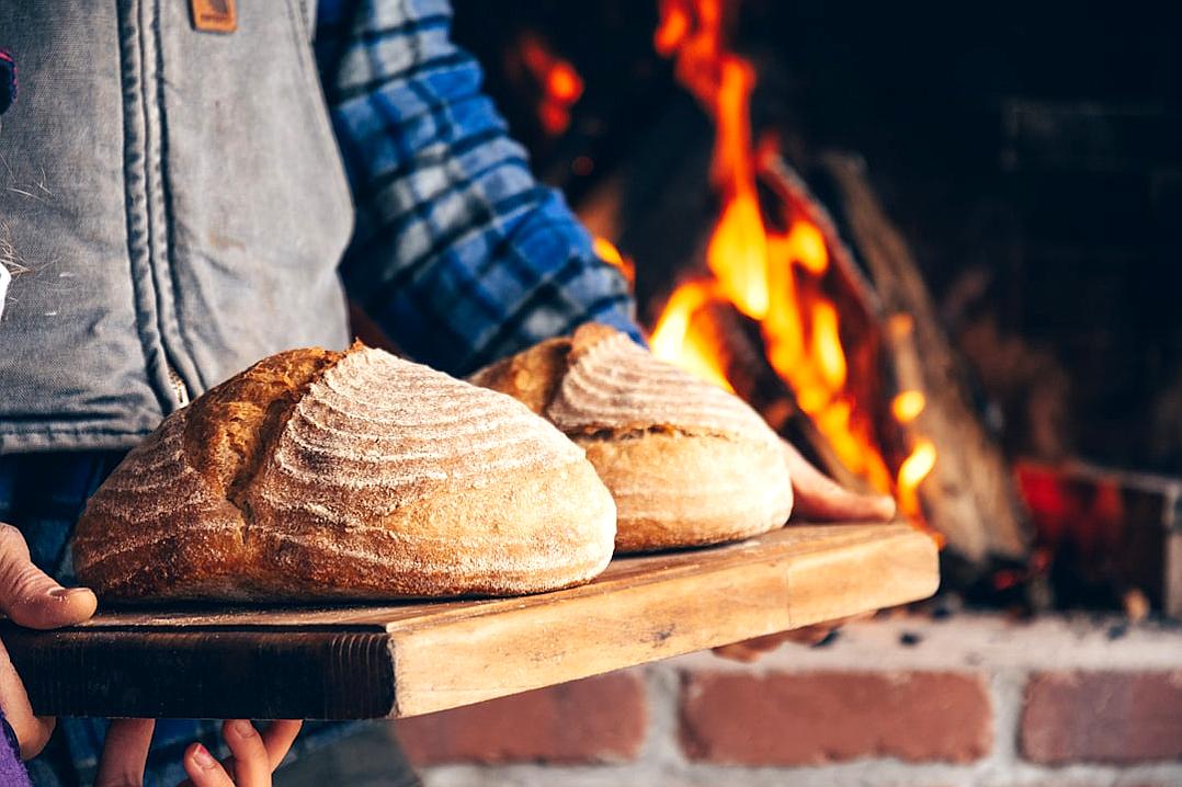 A person holding two sourdough loaves on wooden board, near the fireplace with fire in background, focus is on breads and hands of baker. High resolution photography, insanely detailed, fine details, stock photo, professional color grading, soft shadows, no contrast, clean sharp focus, film lighting