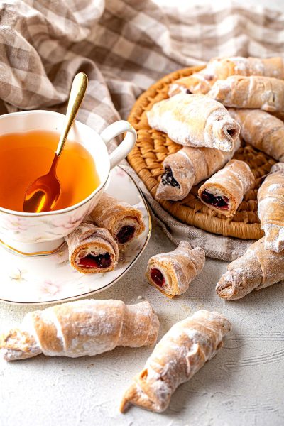 food photography, A pile of small sweet pastry rolls dusted with white sugar and filled with jam on the table next to tea in an elegant cup, golden spoon, soft beige background, light grey fabric, high resolution,