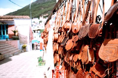 A wall of leather handbags and pouches hanging on the streets in painted buildings, Andalusian style houses with white walls, green mountains behind them, small colorful decorations, the sun is shining brightly. In the style of Christopher Nolan's films, using a Canon EOS camera,