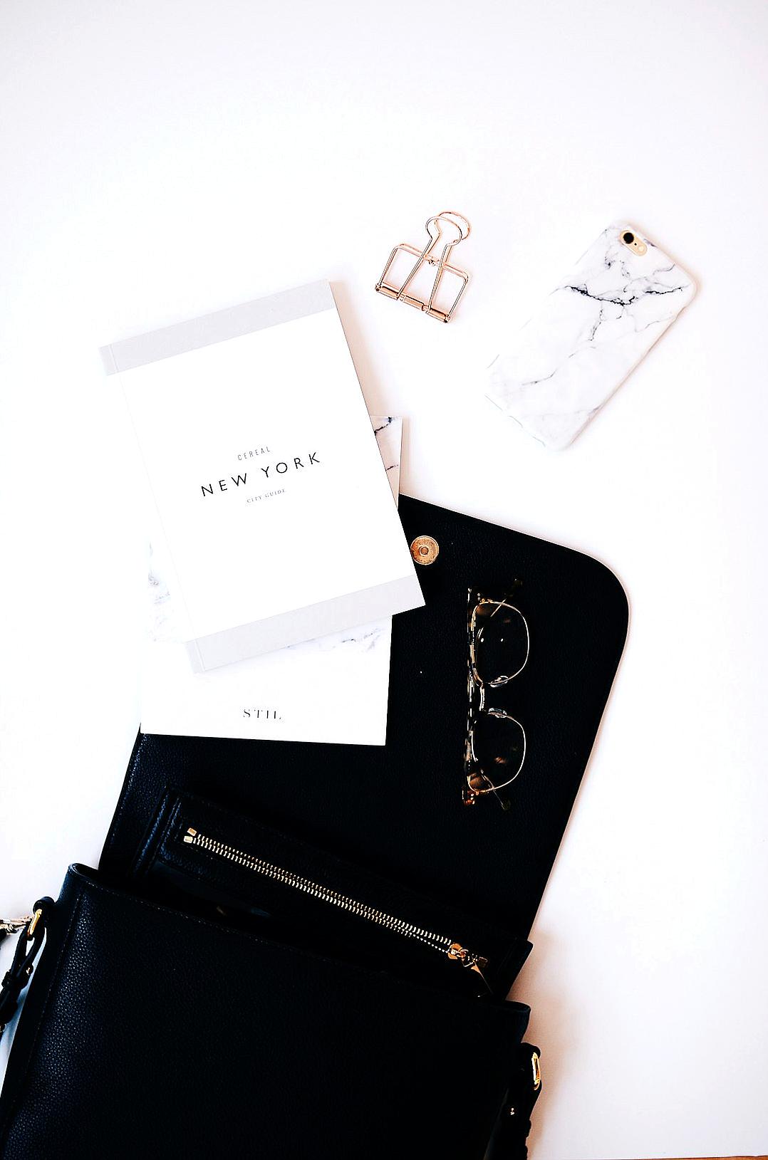 A flat lay of an open black leather bag with glasses and notebook, placed on top of the phone case. The blank card reads “New York” in white text against a marble texture background. A minimalist aesthetic focuses on simplicity and elegance.