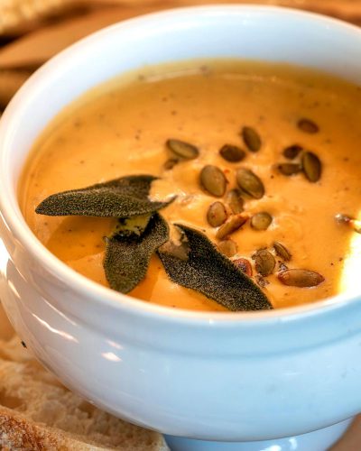 A closeup of the creamy butternut squash soup, with golden pumpkin seeds and fresh sage leaves floating on top in a white bowl, accompanied by slices of baguette bread for side serving. The background is a warm kitchen table, highlighting the rich color and texture of the puree in the style of the cup.