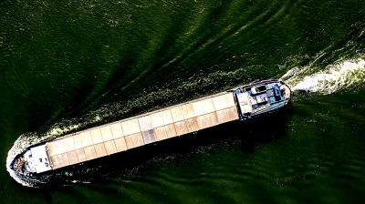 Aerial view of a barge on a river, carrying cargo. A high angle perspective, showcasing the scale and motion of transport in an industrial setting. The water is a dark green with ripples from movement, creating contrast against a bright blue sky. The painting is done in the style of [Claude Monet](https://goo.gl/search?artist%20Claude%20Monet).