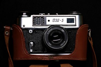 A photo of an old camera with a leather case, front view, on a black background, in a vintage style, of a Leica M3 and Kodak Portra film stock, with studio lighting, a grainy texture, in the style of color chrome photography by Hasselblad X2D, at a high resolution, with cinematic light, hyper realistic, soft shadows, no contrast, with clean sharp focus, a bokeh effect, highly detailed.