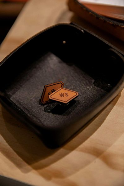 A close-up shot of two leather "WS" patches placed on a black tray, placed next to each other on an empty wooden table. The focus is on capturing intricate details and textures of both elements. Soft lighting highlights their features. The composition showcases the contrast between the dark wood and light brown metal, adding depth to the scene.