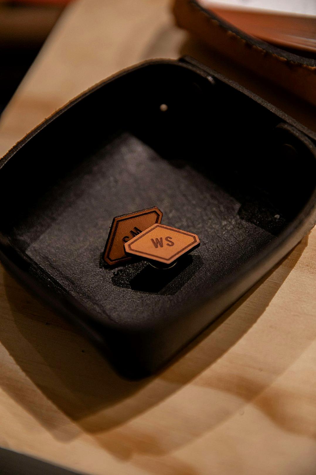 A close-up shot of two leather “WS” patches placed on a black tray, placed next to each other on an empty wooden table. The focus is on capturing intricate details and textures of both elements. Soft lighting highlights their features. The composition showcases the contrast between the dark wood and light brown metal, adding depth to the scene.