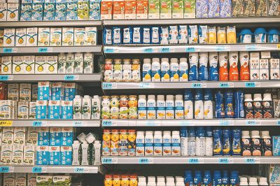 A supermarket shelf full of milk, juice and other small packaging products. The shelves display an array of different bottles, cans and cartons with various colors and shapes. Each product has its own label or logo, indicating the brand name on it. In front is a sign showing some special sales figures. This scene conveys that there's something for everyone in shopping. It shows the variety and diversity the market offers to customers who enjoy exploring new brands and styles in the style of different artists.