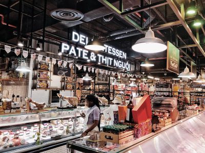 A photo of the inside of an upscale modern grocery store in Vietnam, with a woman working behind one counter and several meat slabs displayed on another counter. Above it all hangs a neon sign that reads "DELI FINE PATRON ORCHESTRA & ENJOY YOUR FOOD." The space is well lit by white lights hanging from the ceiling. In front of each display case there is a glass cover to keep products fresh. It feels clean and spacious, and overall very inviting.