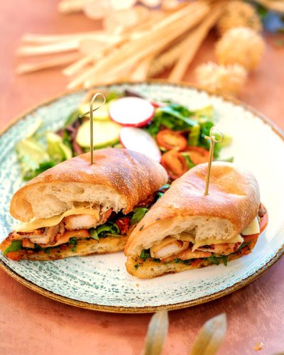 A closeup of two chicken panini sandwiches on an ornate plate, with fresh salad and fries in the background. The sandwich thighs have been sliced into perfect rolls, showcasing their golden brown crusts. A wooden toothpick decorated with leaves stands between each sandwich, adding to its visual appeal. This is a high quality photo that captures every detail of these delicious dishes in the style of a professional food photographer.